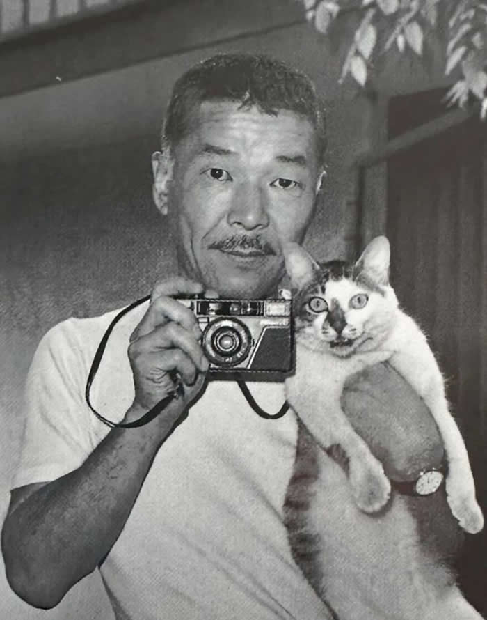 Black-and-white self-portrait of photographer Masahisa Fukase holding a compact camera in one hand and a cat in the other, both facing the viewer.