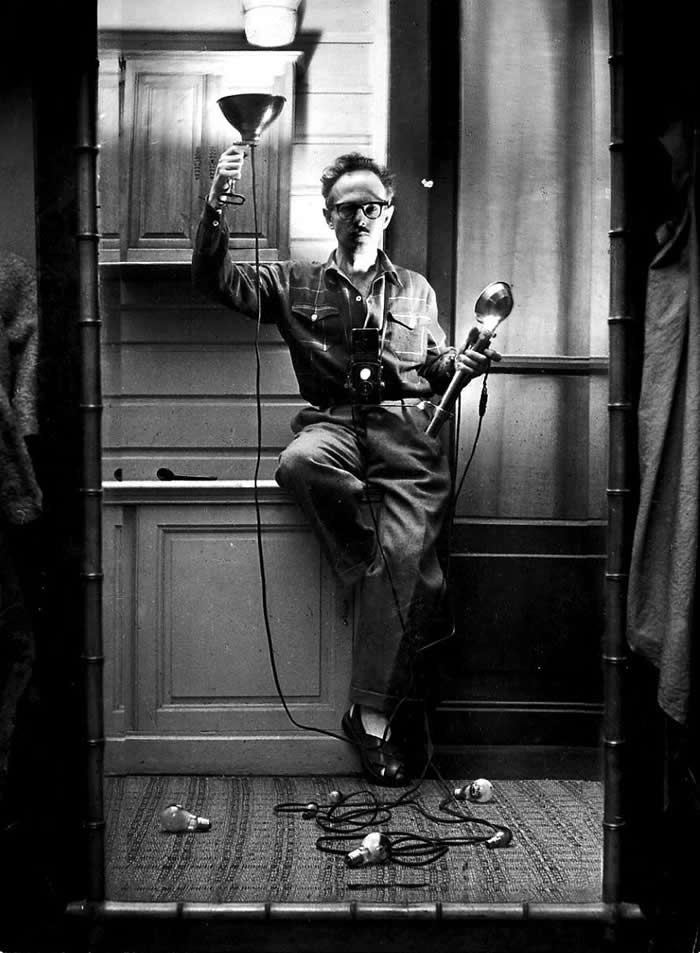 Black-and-white mirror self-portrait of photographer Willy Ronis seated indoors with a camera hanging from his neck, holding two lamps while cords and light bulbs rest on the floor below.
