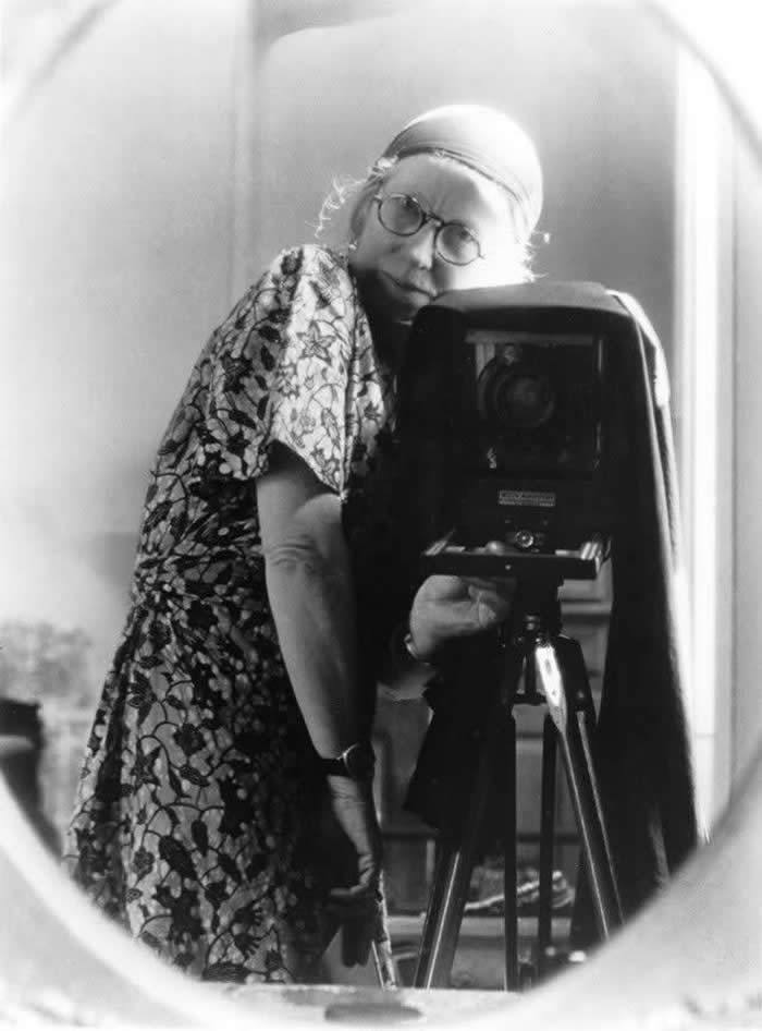 Black-and-white mirror self-portrait of photographer Imogen Cunningham wearing round glasses and patterned clothing, leaning beside a large view camera mounted on a tripod.