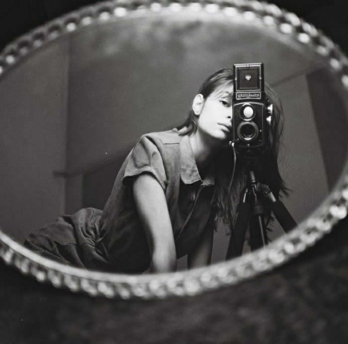 Black-and-white mirror self-portrait of photographer Mirella Cardoso leaning beside a twin-lens reflex camera on a tripod, framed within an oval decorative mirror.