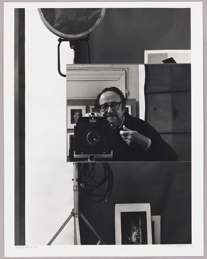 Black-and-white self-portrait of photographer Arnold Newman standing behind a large format camera on a tripod in a studio setting, surrounded by framed photographs and geometric reflections.