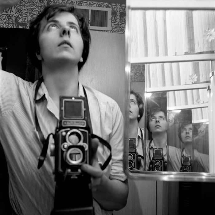 Black-and-white mirror self-portrait of photographer Vivian Maier holding a twin-lens reflex camera, looking upward, with multiple reflections of her face and camera repeating in adjacent mirrors.