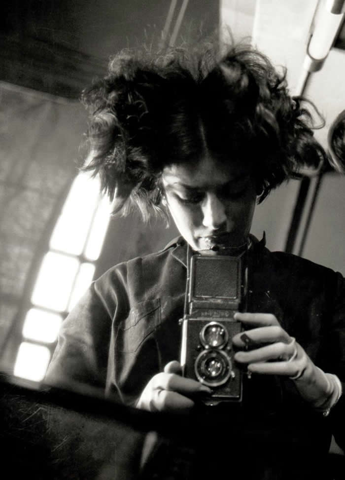 Black-and-white mirror self-portrait of photographer Eva Besny&ouml; looking downward while holding a twin-lens reflex camera, with dramatic light from a nearby window illuminating her face and hair.