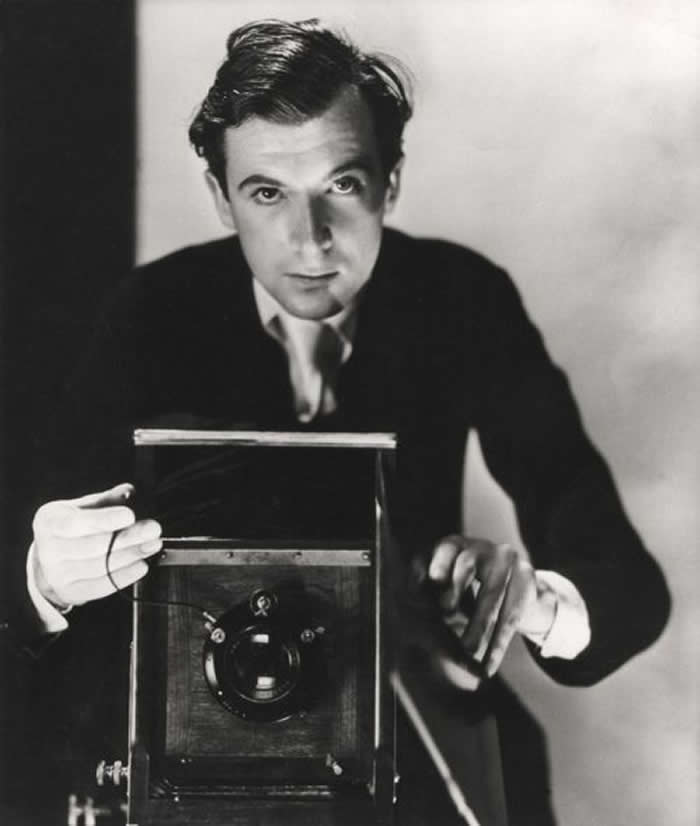 Black-and-white self-portrait of photographer Cecil Beaton standing behind a vintage camera, dressed formally and gazing directly toward the viewer while operating the shutter release.