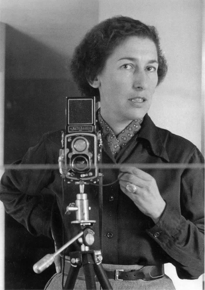 Black-and-white mirror self-portrait of photographer Gis&egrave;le Freund standing beside a twin-lens reflex camera on a tripod, wearing a dark jacket and patterned scarf while looking toward the mirror.