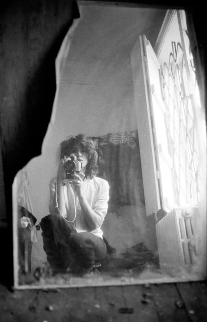 Black-and-white mirror self-portrait of photographer Kate Simon seated indoors holding a camera, reflected in a worn mirror beside an open window with bright light streaming in.