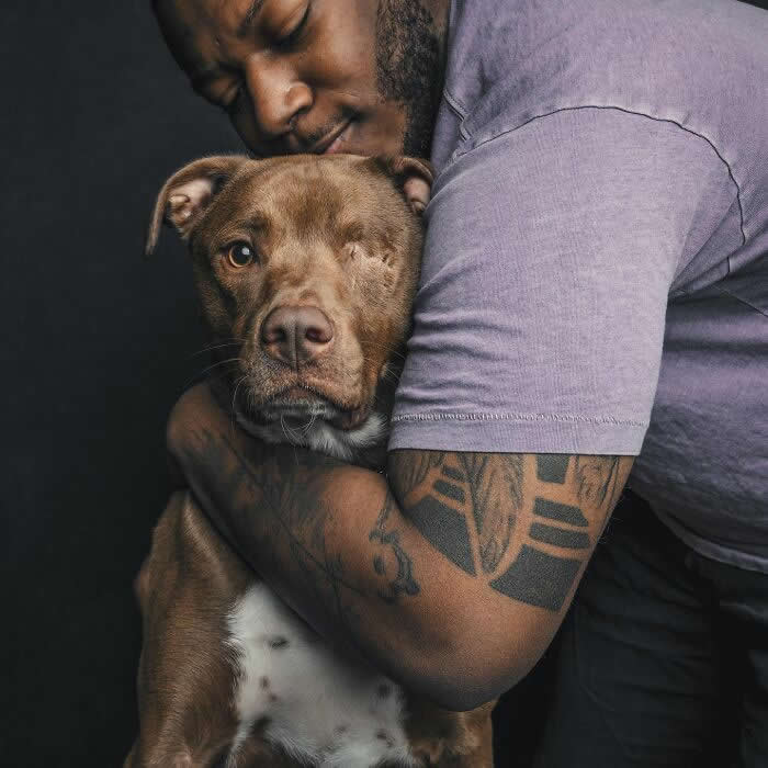 A man with a tattooed arm gently hugs a brown dog with one eye, holding it close in a protective and affectionate embrace against a dark background.