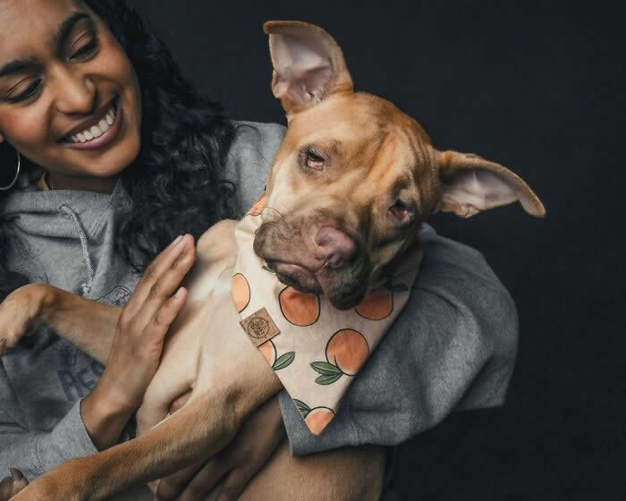 A smiling woman holds a light brown dog wearing a patterned bandana, as the dog tilts its head with a playful and relaxed expression against a dark background.