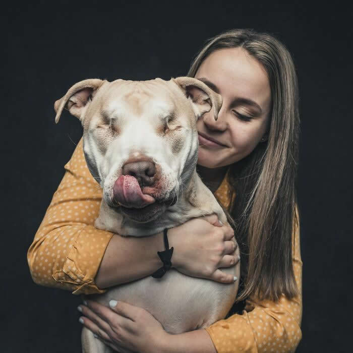 A woman gently hugs a light-colored dog with closed eyes as the dog playfully sticks out its tongue, capturing a warm and affectionate bond.