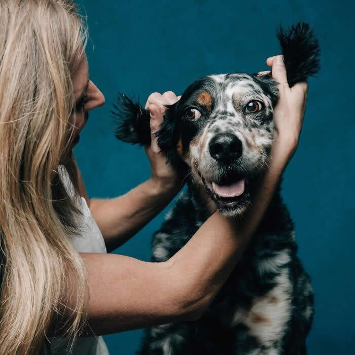 A woman playfully holds the ears of a spotted dog while the dog looks forward with a happy, open-mouthed expression, set against a teal background.