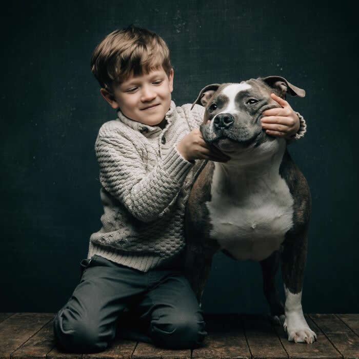 A young boy gently holds and squeezes the cheeks of a calm gray and white dog, capturing a playful and affectionate moment between child and pet.