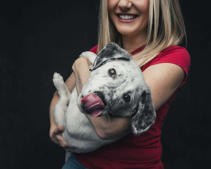 A smiling woman holds a small white and gray spotted puppy in her arms while the puppy sticks out its tongue playfully, creating a joyful and affectionate moment.