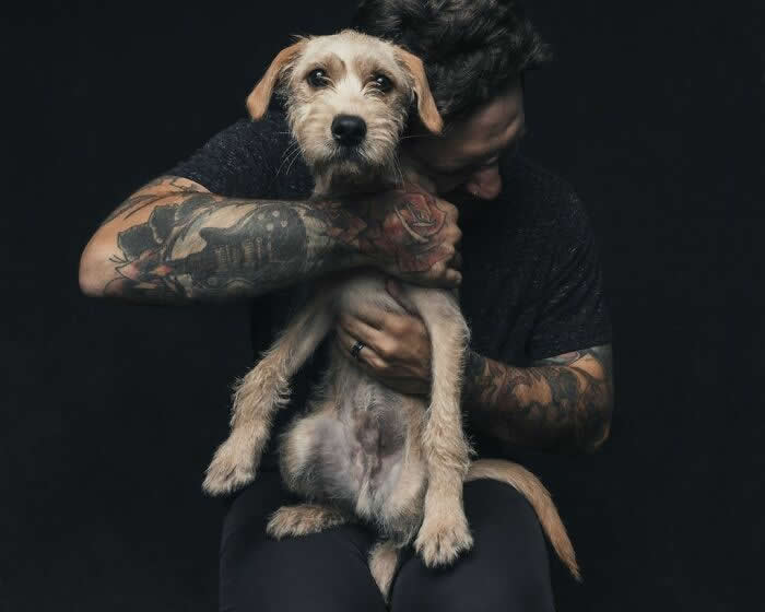 A tattooed man gently holds a small scruffy dog in his arms, embracing it closely while the dog looks forward with a calm and trusting expression against a dark background.