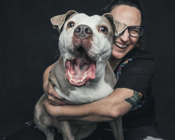 A woman with glasses hugs a large gray and white dog as both appear joyful, with the dog&rsquo;s mouth wide open in a happy expression against a dark background.
