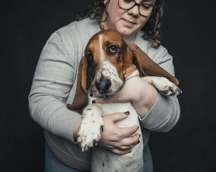 A woman wearing glasses gently cradles a brown and white hound dog with long ears, holding it securely while the dog looks calmly toward the camera against a dark background.