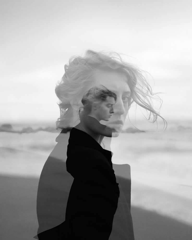 Black and white double exposure portrait of a woman by the sea, blending two overlapping profiles to create a surreal, introspective mood with soft waves in the background.