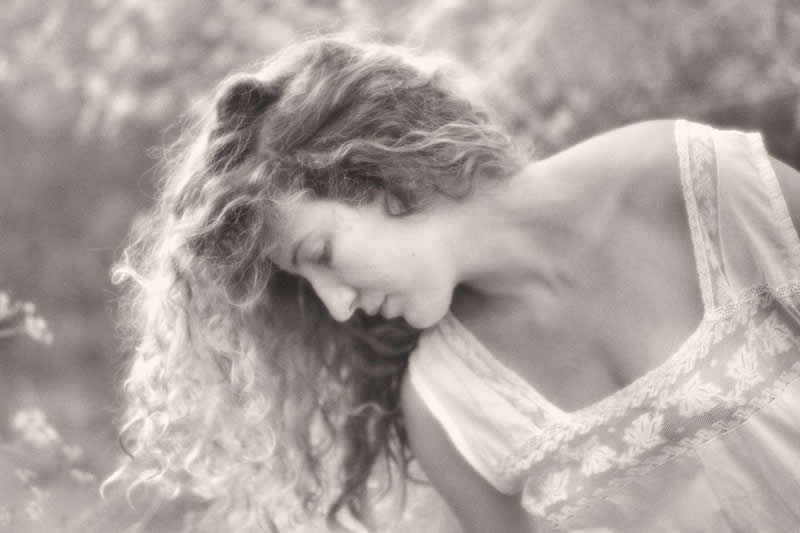 Black and white portrait of a woman with curly hair looking downward, captured in soft natural light with a dreamy, delicate atmosphere and blurred background.