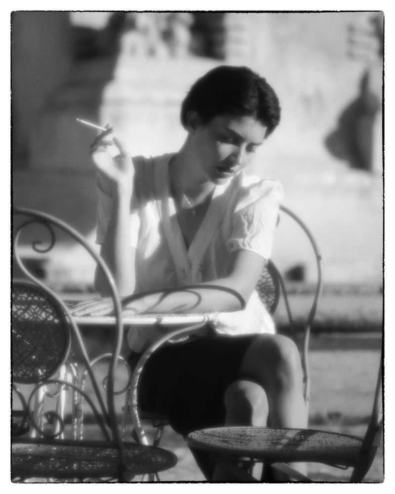 Black and white vintage-style portrait of a woman sitting at an outdoor caf&eacute; table, holding a cigarette, captured in soft light with a dreamy, nostalgic atmosphere.