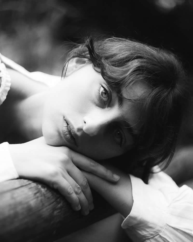 Black and white portrait of a young woman resting her head on her arms, gazing directly at the camera with soft light and a calm, introspective expression.
