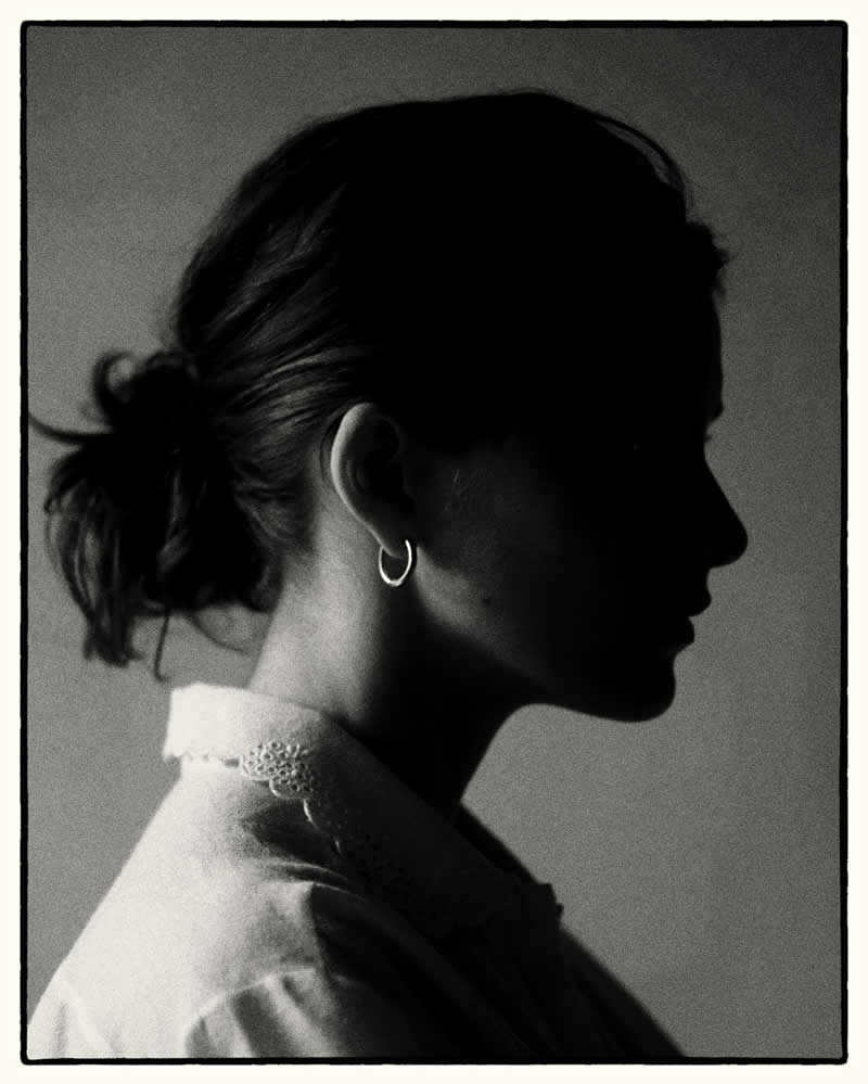 Black and white side-profile silhouette of a woman with tied-back hair and a hoop earring, softly lit against a plain background, creating a minimal and contemplative mood.