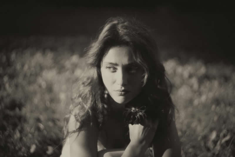 Soft-focus black and white portrait of a woman sitting in a field, looking sideways with a thoughtful expression, surrounded by blurred grass and gentle natural light.