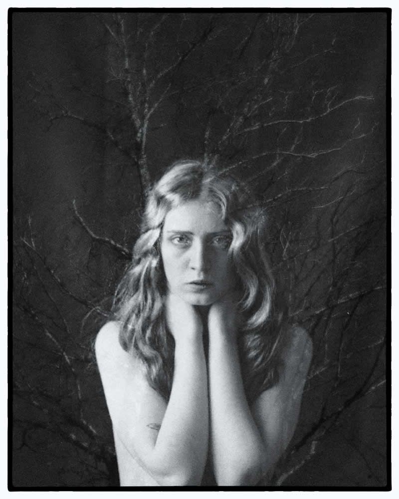 Black and white portrait of a woman with hands near her neck, positioned against a backdrop of bare tree branches that blend with her figure, creating a surreal and moody composition.