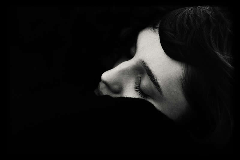 High-contrast black and white close-up of a person&rsquo;s face in profile with eyes closed, partially hidden in shadow, creating a quiet and introspective mood.