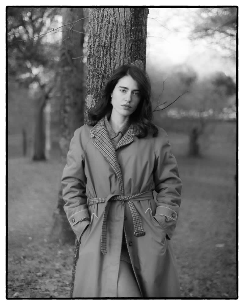 Black and white portrait of a woman wearing a coat, standing against a tree in a park setting, with soft background blur and a calm, contemplative expression.