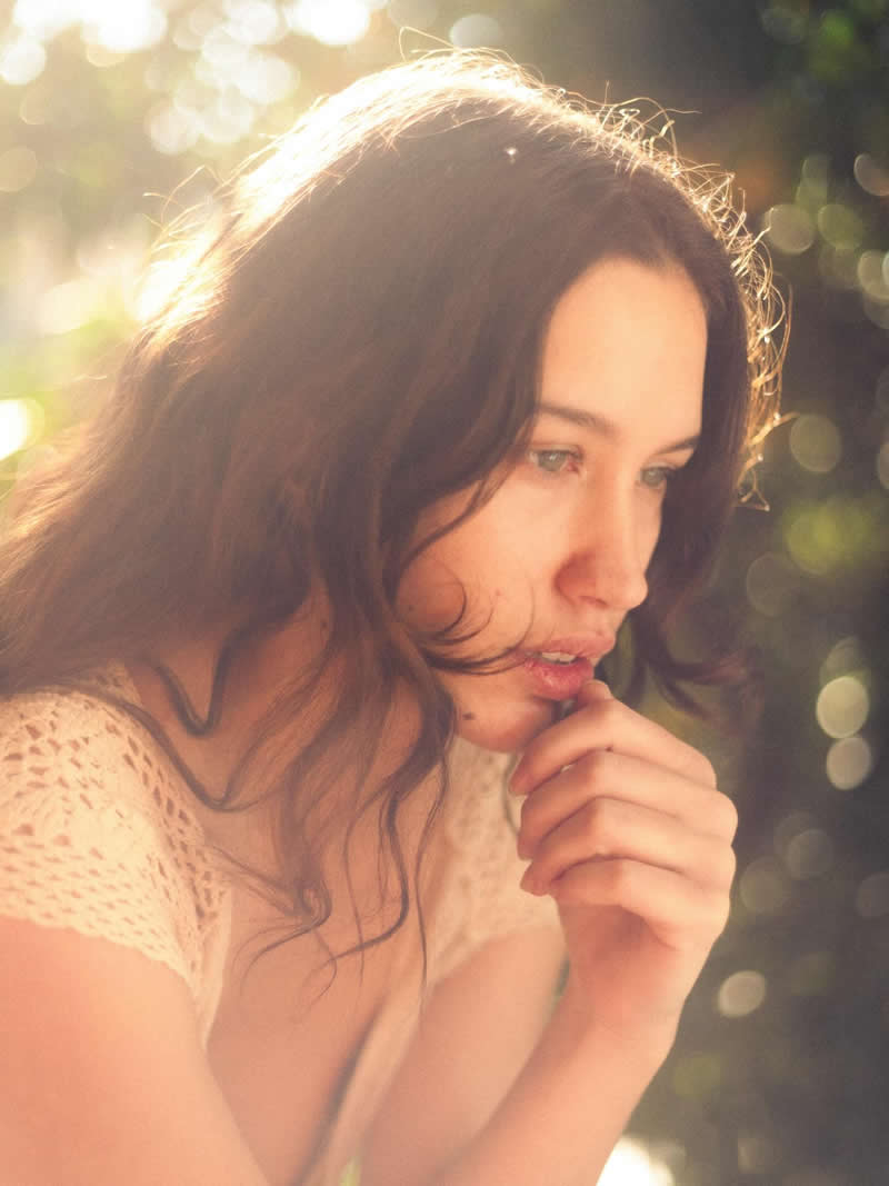 Soft, warm-toned portrait of a woman in natural sunlight, looking thoughtfully to the side with her hand near her lips, surrounded by glowing bokeh and a dreamy atmosphere.
