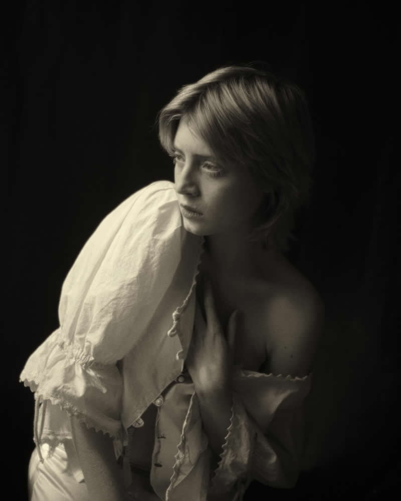Soft, low-key portrait of a woman partially illuminated against a dark background, wearing a delicate blouse, with a gentle, introspective expression and dramatic light falloff.