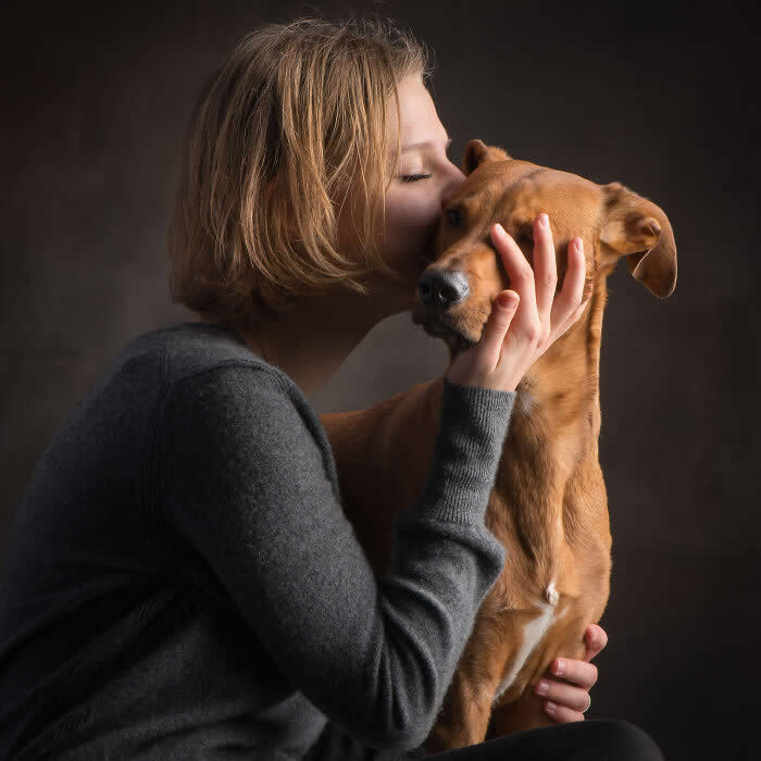 A woman gently kissing a brown dog on the head while holding its face tenderly, captured in soft studio lighting against a dark background, expressing a deep emotional bond and intimacy between human and animal.