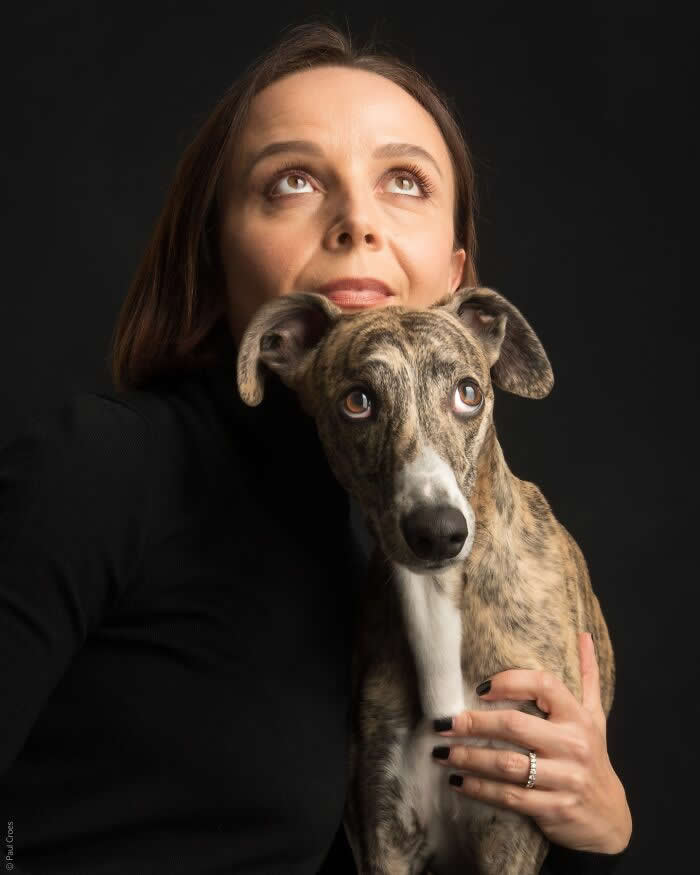A woman gently holds a brindle dog close to her chest, both facing forward under soft studio lighting, with the woman gazing upward and the dog looking at the camera, expressing calm connection and quiet curiosity.