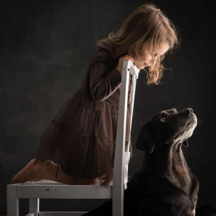 A young girl kneels on a chair, leaning forward as she gazes down at a black dog looking up at her, captured in soft studio lighting, expressing innocence, curiosity, and a gentle human&ndash;animal connection.