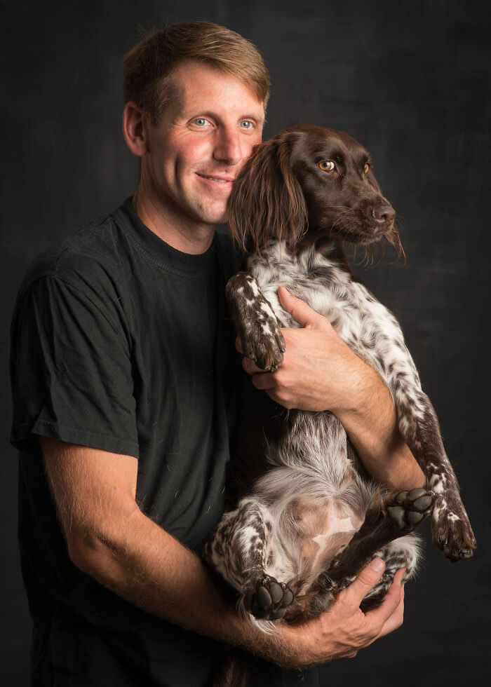 A man gently holds a brown and white dog in his arms, both facing slightly away from the camera, captured in soft studio lighting against a dark background, expressing trust, companionship, and calm connection.