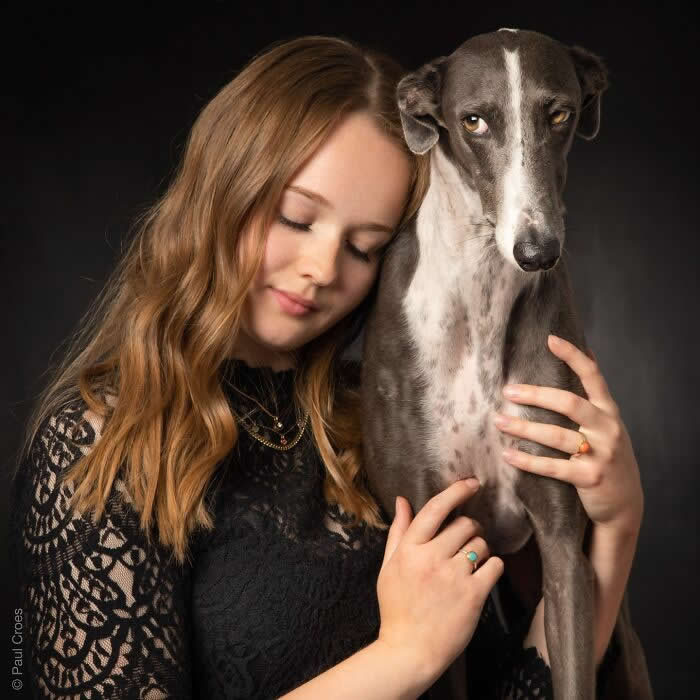 A young woman softly leans her head against a slender grey dog while holding it close, captured in warm studio lighting against a dark background, expressing calm affection, trust, and a quiet human&ndash;animal bond.