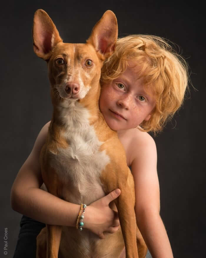 A young child with curly hair gently hugs a slender brown dog, both facing the camera, captured in soft studio lighting against a dark background, expressing innocence, trust, and a strong human&ndash;animal connection.