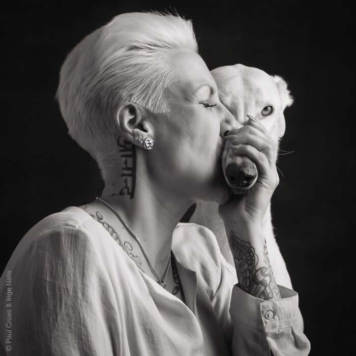 A black-and-white portrait of a woman with short hair gently kissing a white dog&rsquo;s face while holding its muzzle, captured in soft studio lighting, expressing deep affection, trust, and an intimate human&ndash;animal bond.