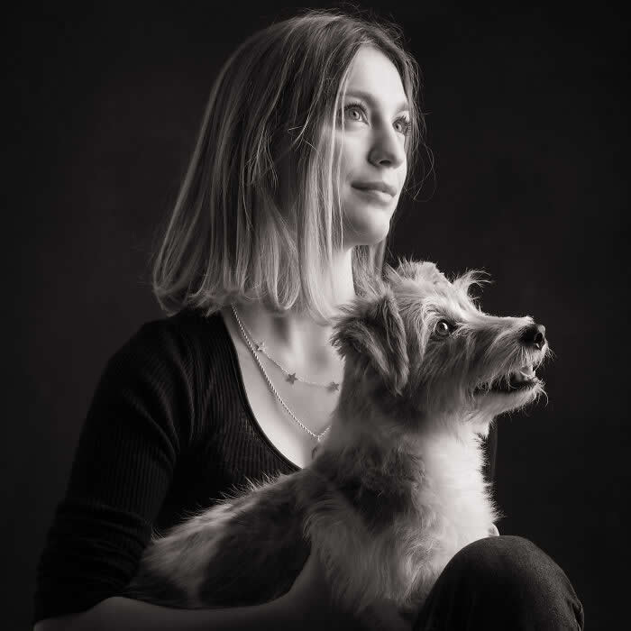 A black-and-white portrait of a woman holding a small dog on her lap, both gazing upward in the same direction, softly lit against a dark background, expressing hope, connection, and shared emotion.