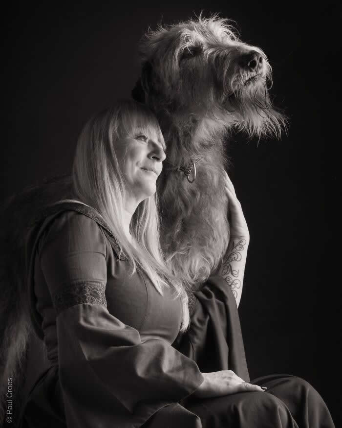 A black-and-white portrait of a woman sitting beside a large, long-haired dog, both gazing upward, softly lit against a dark background, expressing companionship, calm strength, and a shared sense of hope.