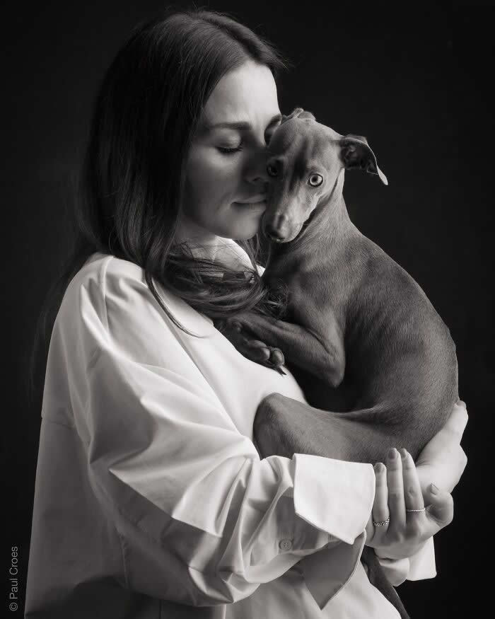 A black-and-white portrait of a woman gently cradling a small dog against her chest, eyes closed in a tender moment, captured in soft studio lighting, expressing intimacy, trust, and deep emotional connection.