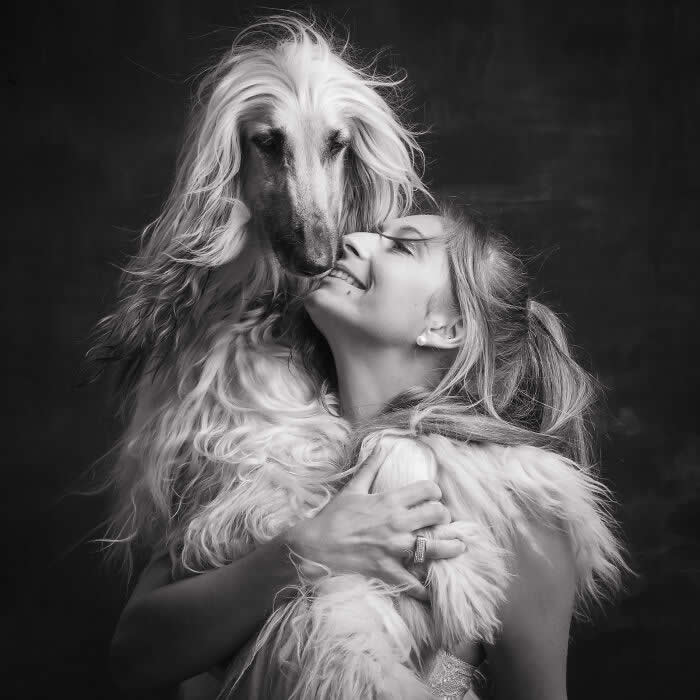 Black-and-white portrait of a woman smiling with eyes closed as she gently embraces a long-haired Afghan hound, their faces touching, conveying deep affection, elegance, and an intimate human-animal bond.