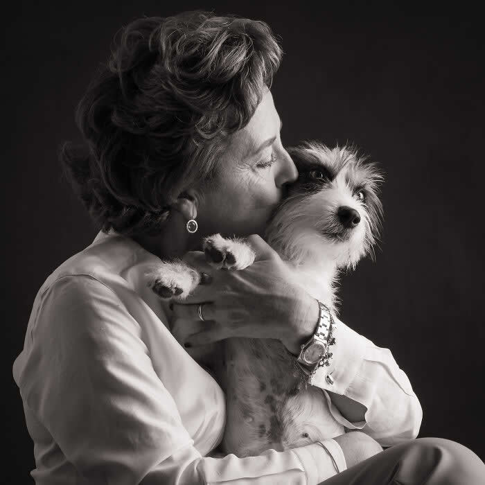 Black-and-white portrait of a woman gently kissing a small dog on the head while holding it close, the dog gazing outward, expressing tenderness, trust, and a deep human-animal bond.
