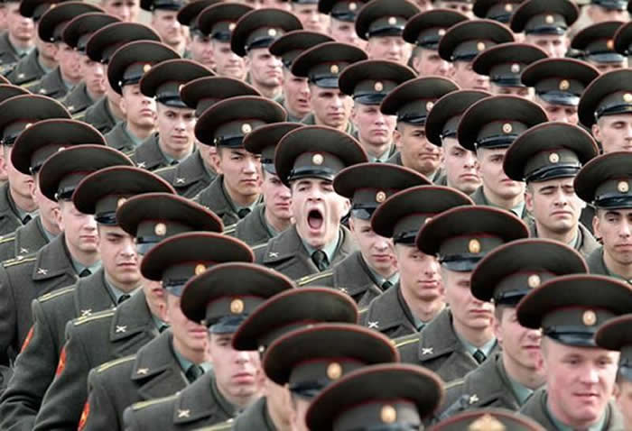 A high-angle shot of a large group of soldiers in olive green uniforms and peaked caps standing in tight formation. In the center of the crowd, one soldier has his mouth wide open in a large, mid-action yawn.