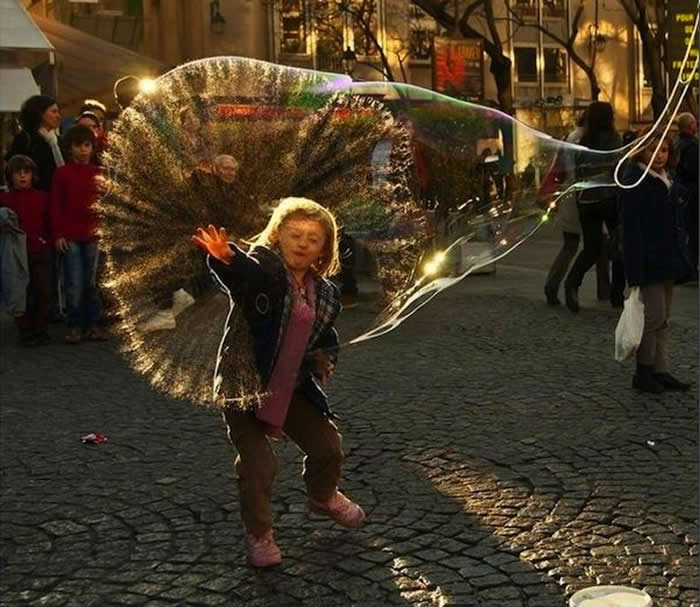 A perfectly timed photo of a young girl on a cobblestone street as a large soap bubble bursts around her head. The thin, iridescent film of the bubble is caught mid-shatter, completely encircling her face and hair like a clear mask.