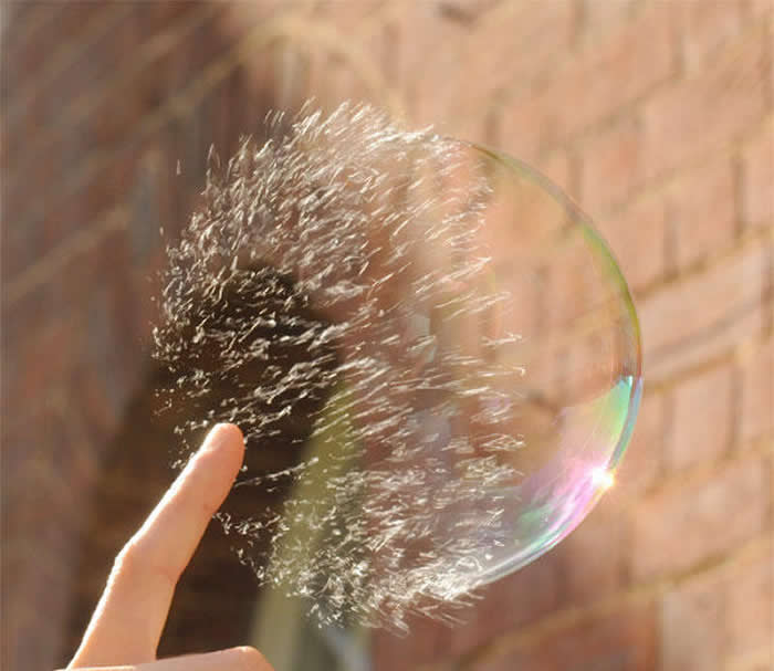 A high-speed, perfectly timed photo of a finger touching a large soap bubble against a brick wall. The right side of the bubble is still intact and shimmering, while the left side is disintegrating into a mist of droplets.