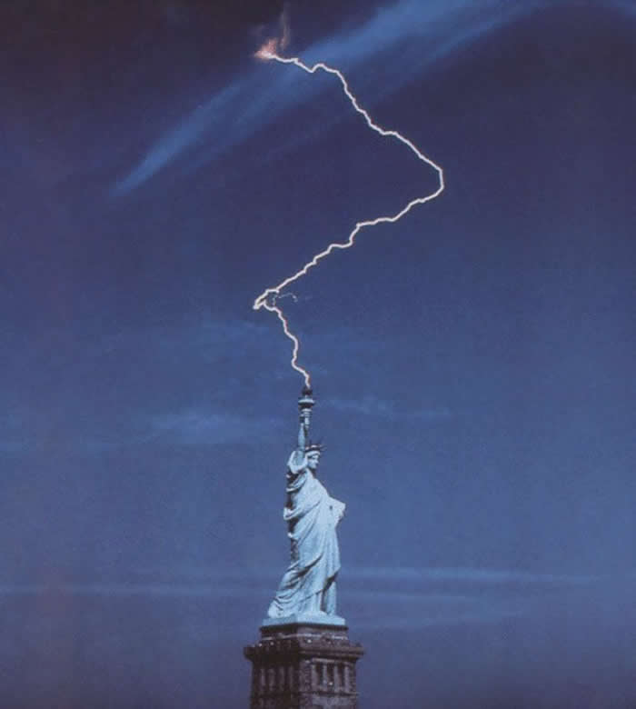 A dramatic, perfectly timed night photograph of a single lightning bolt striking the torch of the Statue of Liberty. The sky is a deep blue, and the lightning creates a bright, zigzagging line from the clouds to the monument's torch.