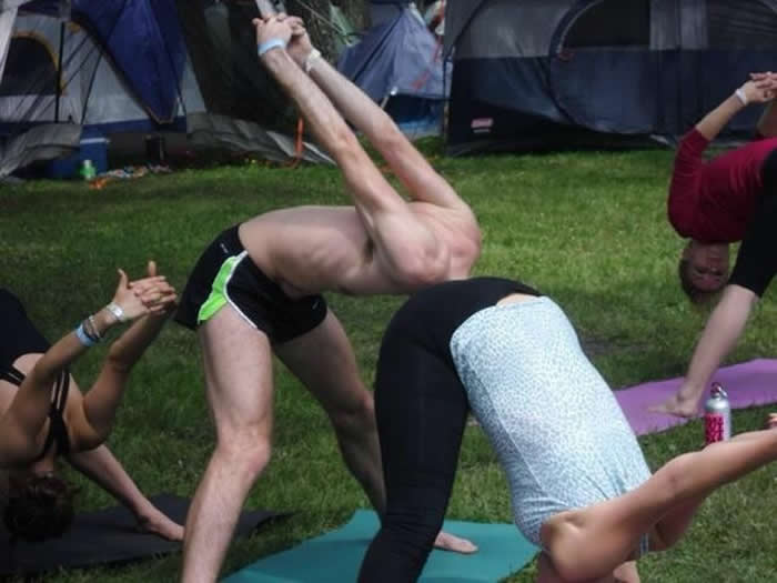 An optical illusion photo of two people practicing yoga outdoors. A shirtless man leans forward and a person in a blue top leans back, with their bodies positioned so they appear to be one continuous, strangely shaped person.