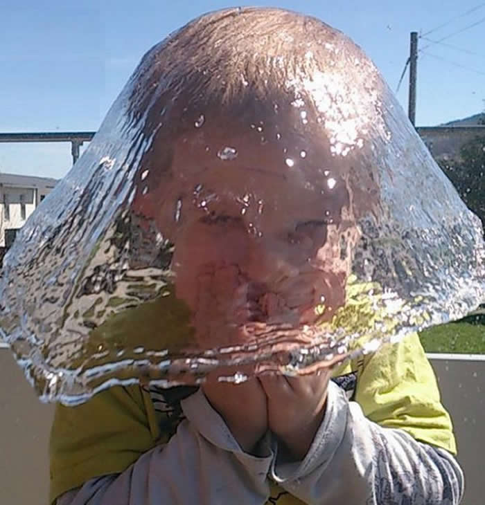 A perfectly timed high-speed photo of a young boy as a water balloon bursts over his head. The water forms a clear, dome-like sheet that perfectly covers his face and hair, creating the illusion of a liquid helmet.