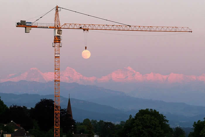 A perfectly timed photograph of a yellow construction crane against a dusky sky. The full moon is positioned exactly beneath the crane's hoist, making it look as though the machine is lifting the moon over a mountain range.