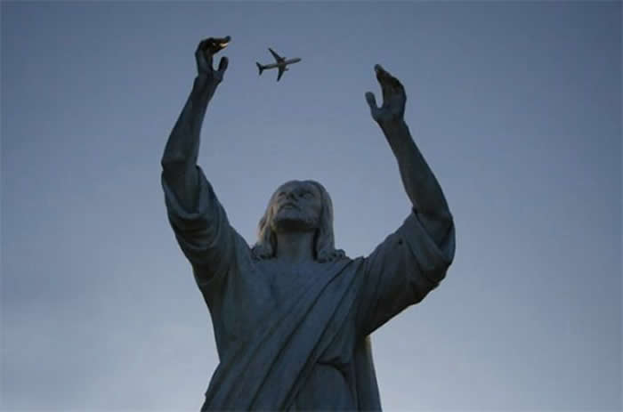 A perfectly timed forced-perspective photo of a stone statue of Jesus with his arms raised and palms open. A small airplane in the sky is positioned exactly between his hands, making it look as though the statue is catching the plane.